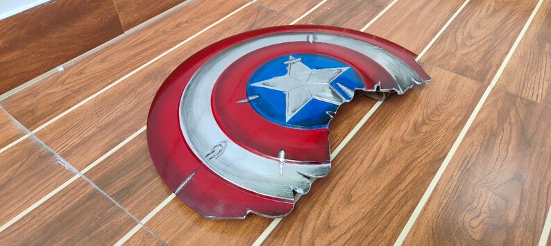 May include: A distressed, half-circle shield in red, white, and blue, featuring a central blue circle with a white star. The shield has a metallic silver trim and a battle-worn appearance, with visible damage along the edges.