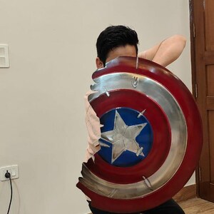 May include: A Captain America shield prop, featuring a red, white, and blue color scheme with a silver metallic rim. The shield has a star in the center and appears to be damaged. The person is wearing black pants.