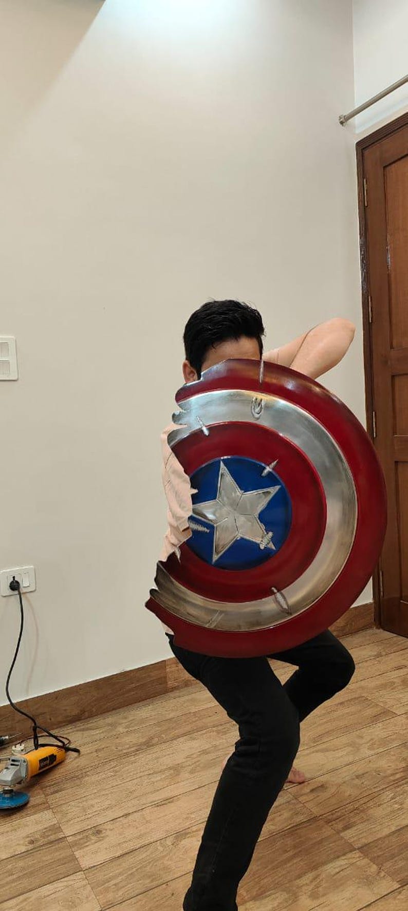 May include: A Captain America shield prop, featuring a red, white, and blue color scheme with a silver metallic rim. The shield has a star in the center and appears to be damaged. The person is wearing black pants.