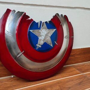 May include: A distressed, red, white, and blue shield with a silver trim and a white star in the center. The shield has a battle-worn appearance with a large section missing from the top. The shield is a prop or collectible.