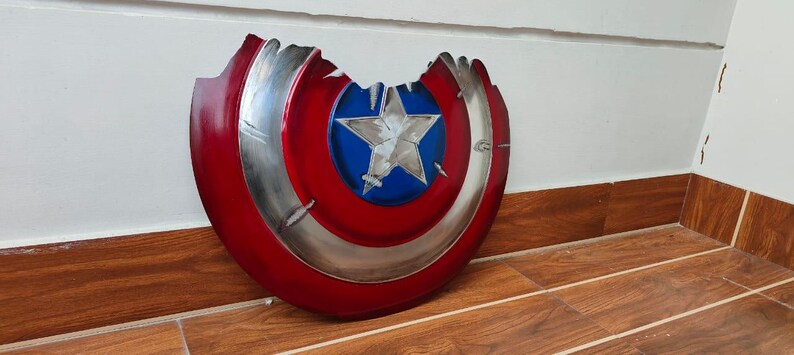 May include: A distressed, red, white, and blue shield with a silver trim and a white star in the center. The shield has a battle-worn appearance with a large section missing from the top. The shield is a prop or collectible.