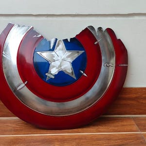 May include: A distressed Captain America shield, featuring a red and silver design with a blue star in the center. The shield has a battle-worn appearance with a section missing, showcasing a weathered aesthetic. The shield is a prop or collectible.