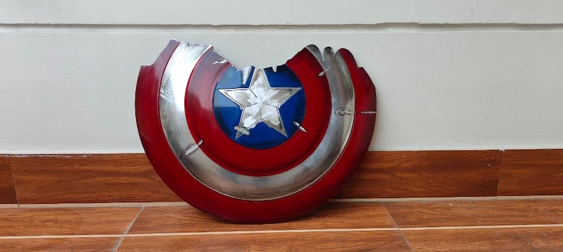 May include: A distressed Captain America shield, featuring a red and silver design with a blue star in the center. The shield has a battle-worn appearance with a section missing, showcasing a weathered aesthetic. The shield is a prop or collectible.