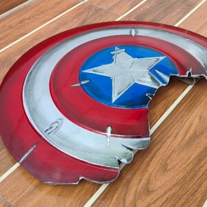 May include: A distressed, half-circle shield in red, white, and blue, featuring a central blue circle with a white star. The shield has a metallic silver trim and a battle-worn appearance, with visible damage along the edge.