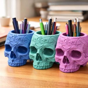 Peut inclure: Trois porte-stylos en forme de crâne colorés en bleu, vert d'eau et rose, chacun rempli de stylos et de crayons. Les supports sont en matériau texturé et reposent sur une surface en bois. Un organisateur de bureau unique.