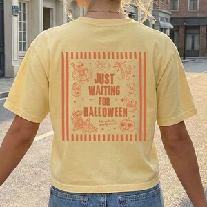 May include: Yellow t-shirt with a Halloween-themed graphic on the back. The graphic includes the text "JUST WAITING FOR HALLOWEEN" with orange illustrations of a skeleton, palm tree, and pumpkin. The shirt is a light yellow color.