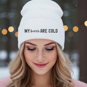 May include: A white beanie with the text "MY bones ARE COLD" in black. The beanie is worn by a person with blonde hair and pink clothing. The background is blurred with bokeh lights.