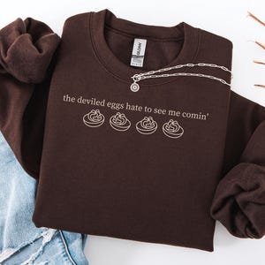 May include: A dark brown sweatshirt with the phrase "the deviled eggs hate to see me comin'" and a graphic of deviled eggs in white. A silver necklace is draped across the sweatshirt. Light blue ripped jeans are visible in the background.