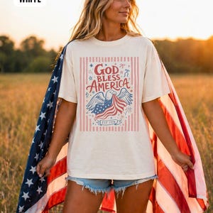 May include: A white t-shirt with a patriotic design featuring an eagle, American flag, and the text "God Bless America." The shirt has a vintage look with red and blue accents and vertical stripes. The word "WHITE" is in the top left corner.