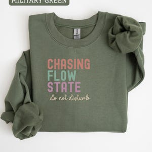 May include: Military green sweatshirt with the text "CHASING FLOW STATE do not disturb" in pastel colors. The long-sleeved crewneck top is made of a soft material and is suitable for casual wear.