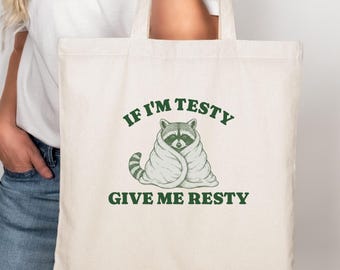 May include: Cream-colored tote bag with a green graphic of a raccoon wrapped in a blanket. The text "IF I'M TESTY GIVE ME RESTY" is printed above and below the raccoon illustration. The bag has two long handles.