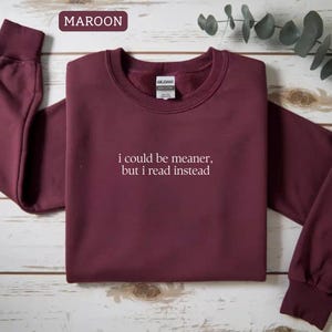 May include: Maroon sweatshirt with the text "i could be meaner, but i read instead" in white. The sweatshirt has a crew neck and long sleeves with ribbed cuffs. The word "MAROON" is in a rectangular box in the upper left corner.