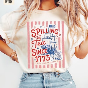 May include: Ivory-colored t-shirt with a graphic that reads "Spilling the Tea Since 1773" with a ship illustration. The design includes red and blue accents, stars, and vertical pink stripes. The text "250 Years of Freedom" is also included.