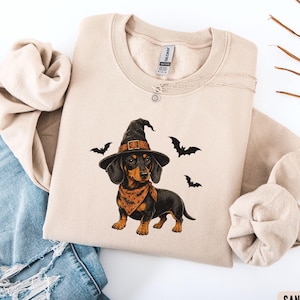 May include: A sand-colored crewneck sweatshirt featuring a graphic of a black and tan dachshund wearing a pointed witch hat and an orange bandana with a bat pattern. Three black bats are illustrated flying around the dog on the front.