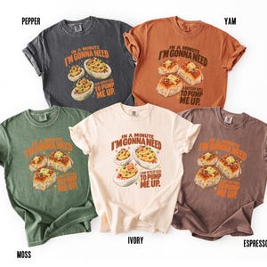 May include: Five short-sleeve t-shirts in charcoal, orange, moss green, ivory, and espresso. Each shirt features a graphic print of food items with the text "IN A MINUTE I'M GONNA NEED TO PUMP ME UP" and specific food labels like deviled eggs or buttery rolls.