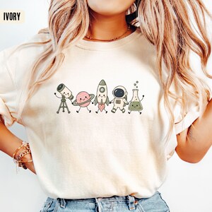 May include: An ivory short-sleeve t-shirt featuring a graphic print of five cute, anthropomorphic space-themed characters holding hands: a telescope, a planet with rings, a rocket ship, an astronaut, and a laboratory flask with green liquid.