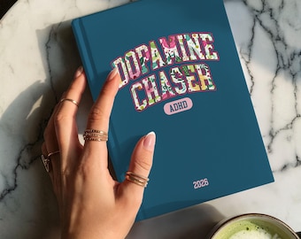 May include: A teal-colored notebook with the words "Dopamine Chaser" in a floral design. The word "ADHD" is below the title, and the year "2026" is at the bottom. The notebook is on a marble surface.