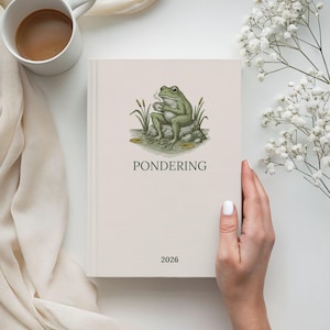 May include: A cream-colored journal with the word "PONDERING" and the year "2026" printed on the cover. The cover features a green frog illustration. A white mug of coffee and flowers are in the background.
