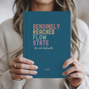 May include: A teal-colored notebook with the words "GENUINELY REACHED FLOW STATE do not disturb" in colorful text. The year "2026" is printed at the bottom. The notebook is held by a person.