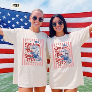 May include: Ivory-colored t-shirts with a graphic that reads "Spilling the Tea Since 1773" and a ship illustration. The shirts are worn in front of an American flag. The text also includes "250 Years of Freedom".