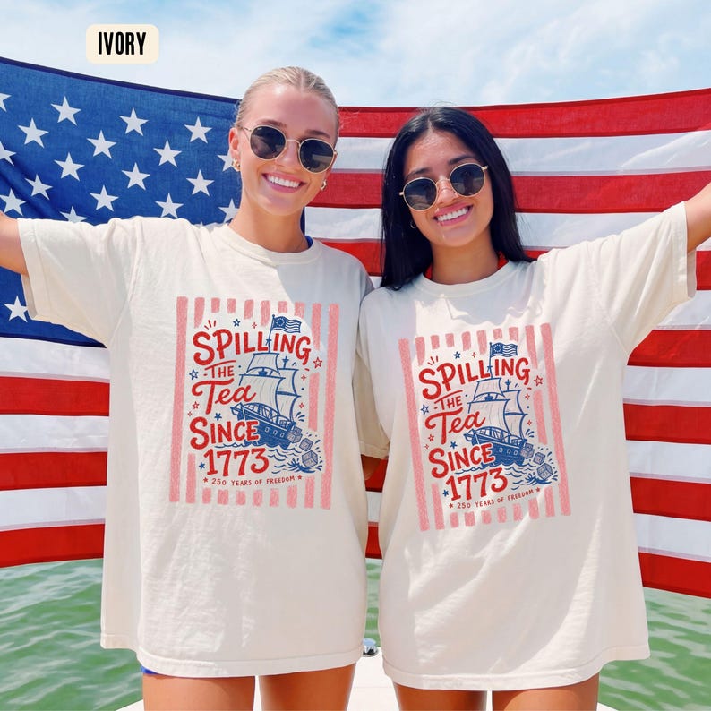May include: Ivory-colored t-shirts with a graphic that reads "Spilling the Tea Since 1773" and a ship illustration. The shirts are worn in front of an American flag. The text also includes "250 Years of Freedom".