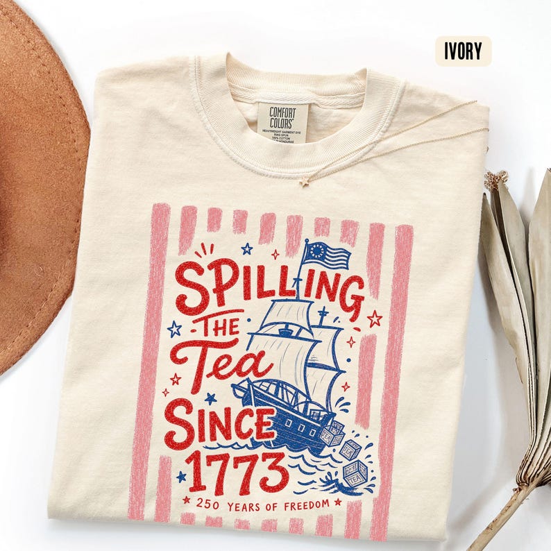 May include: Ivory-colored t-shirt with a graphic that reads "Spilling the Tea Since 1773." The design features a ship, stars, and red and pink stripes. The shirt is made of a soft material and has a crew neck.