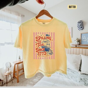 May include: A yellow t-shirt with a graphic that reads "Spilling the Tea Since 1773" with a ship illustration. The design is in red, blue, and white, set against pink vertical stripes. The shirt is hanging on a wooden hanger.