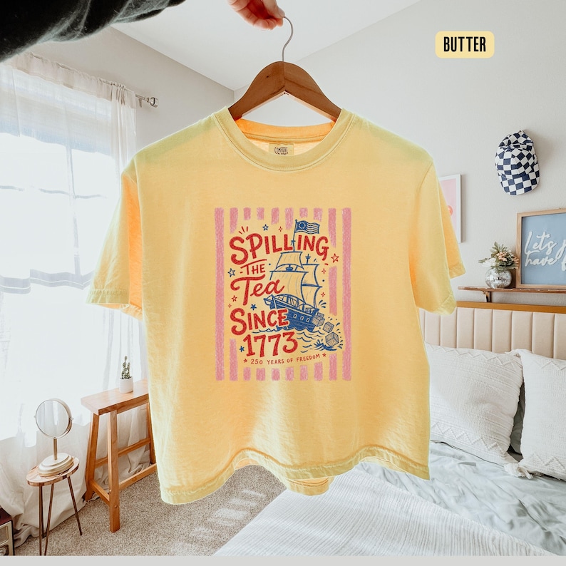 May include: A yellow t-shirt with a graphic that reads "Spilling the Tea Since 1773" with a ship illustration. The design is in red, blue, and white, set against pink vertical stripes. The shirt is hanging on a wooden hanger.