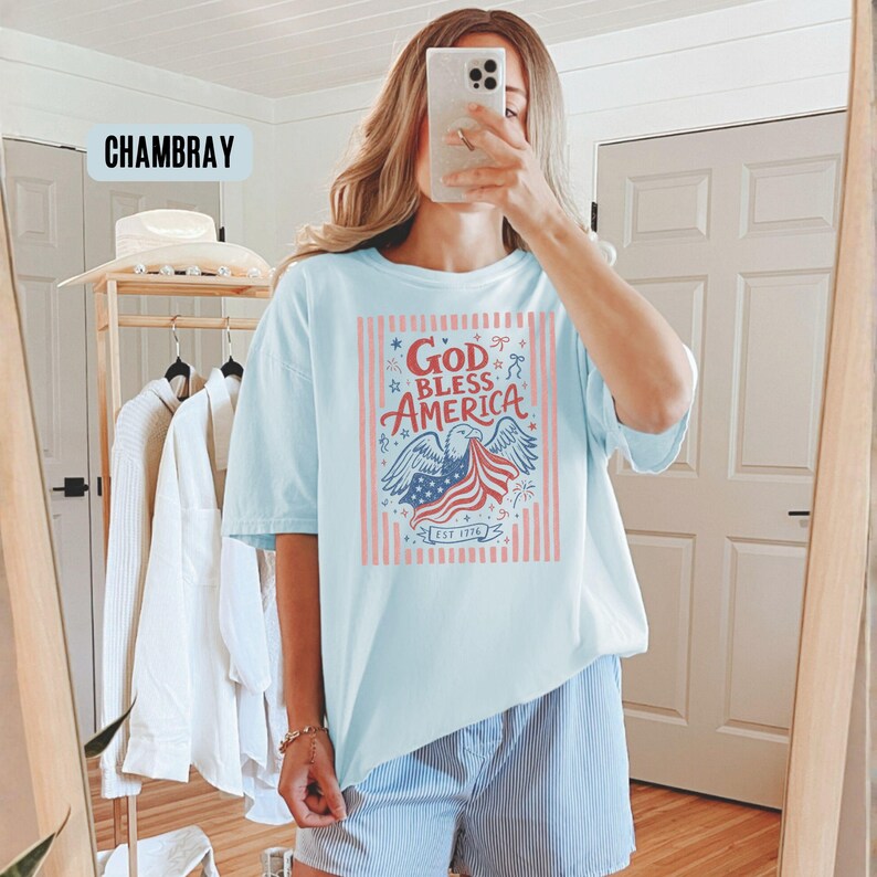 May include: Light blue t-shirt with a patriotic graphic. The design features an eagle, the American flag, and the text "God Bless America" in red, white, and blue. The shirt has short sleeves and a relaxed fit.