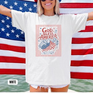 May include: White t-shirt with a patriotic design. The graphic features an eagle holding a waving American flag, with the words "God Bless America" and "Est 1776" in a decorative font. The shirt is displayed in front of an American flag.