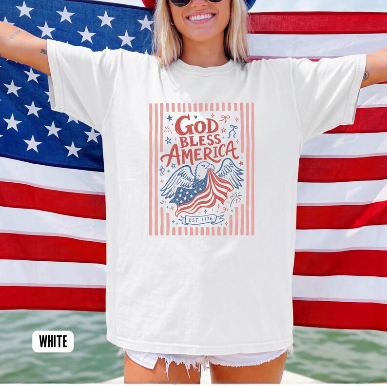 May include: White t-shirt with a patriotic design. The graphic features an eagle holding a waving American flag, with the words "God Bless America" and "Est 1776" in a decorative font. The shirt is displayed in front of an American flag.