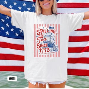 May include: White t-shirt with a patriotic design featuring a ship, the text "Spilling the Tea Since 1773," and the phrase "250 Years of Freedom." The design is in red, white, and blue, with a vertical striped background.