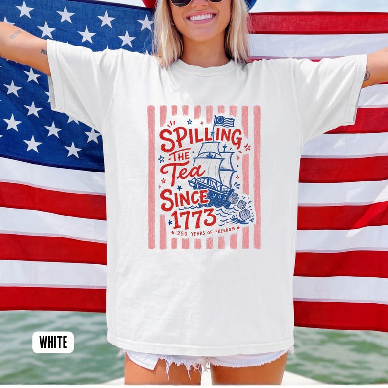 May include: White t-shirt with a patriotic design featuring a ship, the text "Spilling the Tea Since 1773," and the phrase "250 Years of Freedom." The design is in red, white, and blue, with a vertical striped background.