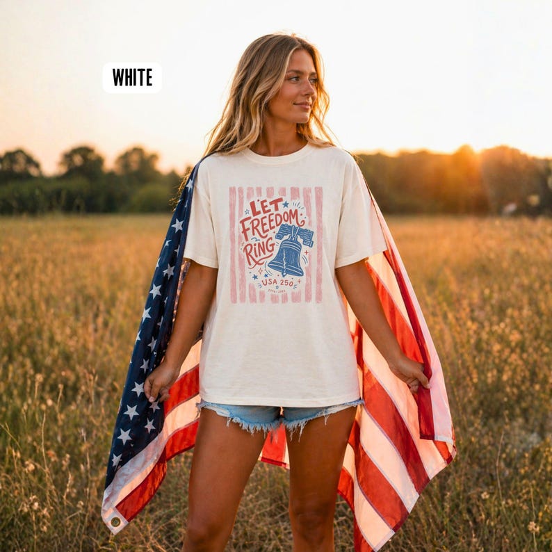 K&ouml;nnte beinhalten: Cremefarbenes T-Shirt mit dem Text "Let Freedom Ring" und einer Glockengrafik. Das Shirt wird mit einer US-Flagge &uuml;ber den Schultern getragen. Der Text "USA 250" befindet sich unter der Glockengrafik. Das Wort "WHITE" steht oben links.