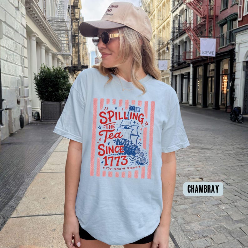 May include: Light blue t-shirt with the text "Spilling the Tea Since 1773" and a graphic of a ship. The design includes red and white stripes and stars. The shirt is a casual style.