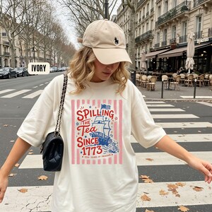 May include: Ivory-colored t-shirt with a graphic that reads "Spilling the Tea Since 1773" featuring a ship illustration. The shirt is paired with a beige baseball cap with a black logo and a black purse. The shirt design includes red, white, and blue colors.