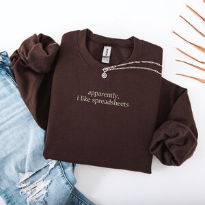 May include: A dark brown sweatshirt with the embroidered text "apparently, i like spreadsheets." A silver necklace with a circular pendant rests on the sweatshirt. Light blue ripped jeans are visible on the left side of the image.