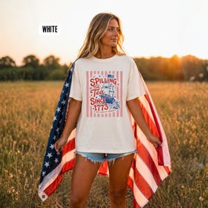 May include: Cream-colored t-shirt with the text "Spilling the Tea Since 1773" and a ship graphic. The shirt is worn with denim shorts and an American flag. The shirt has a relaxed fit and is suitable for casual wear.