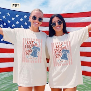 K&ouml;nnte beinhalten: Elfenbeinfarbene T-Shirts mit dem Text "Let Freedom Ring" und einer Glockengrafik. Die Shirts werden vor einer amerikanischen Flagge getragen. Der Text "USA 250" ist ebenfalls auf den Shirts aufgedruckt.