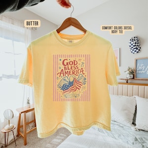 May include: A yellow Comfort Colors boxy tee shirt with the words "God Bless America" and an eagle graphic holding an American flag. The shirt has short sleeves and a crew neck. The text "EST 1776" is also visible.
