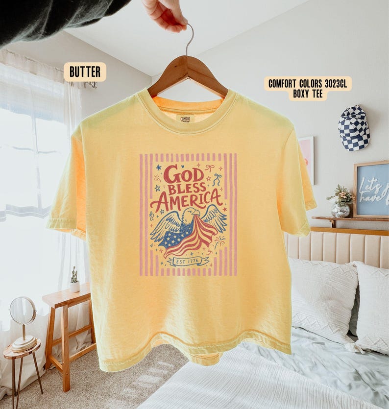 May include: A yellow Comfort Colors boxy tee shirt with the words "God Bless America" and an eagle graphic holding an American flag. The shirt has short sleeves and a crew neck. The text "EST 1776" is also visible.