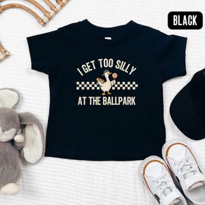May include: Black toddler t-shirt with the text "I GET TOO SILLY AT THE BALLPARK" and a cartoon goose holding a lollipop. The shirt is paired with a black cap and white sneakers with brown accents. A gray stuffed bunny and a woven wall decor are also in the image.