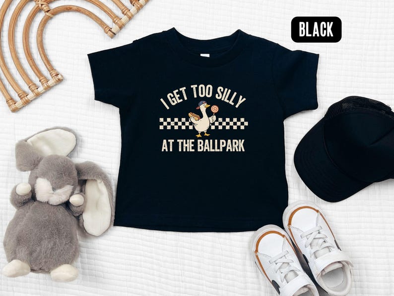 May include: Black toddler t-shirt with the text "I GET TOO SILLY AT THE BALLPARK" and a cartoon goose holding a lollipop. The shirt is paired with a black cap and white sneakers with brown accents. A gray stuffed bunny and a woven wall decor are also in the image.