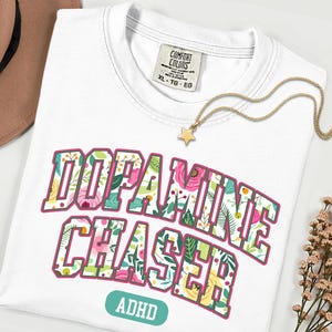 Pode incluir: Camiseta branca com as palavras "DOPAMINE CHASER" em um design floral, com "ADHD" abaixo. Um colar com pingente de estrela dourada e um chap&eacute;u marrom tamb&eacute;m s&atilde;o vis&iacute;veis.