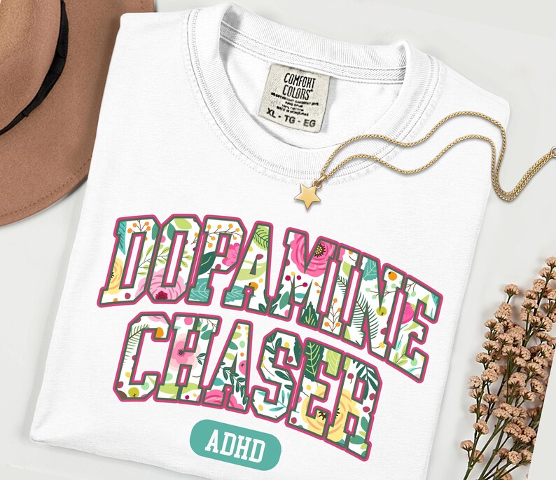 Pode incluir: Camiseta branca com as palavras "DOPAMINE CHASER" em um design floral, com "ADHD" abaixo. Um colar com pingente de estrela dourada e um chap&eacute;u marrom tamb&eacute;m s&atilde;o vis&iacute;veis.