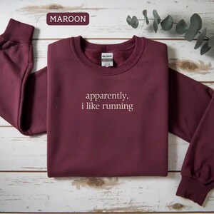 Może przedstawiać: Bordowa bluza z okrągłym dekoltem z białym napisem "apparently, i like running". Bluza ma długie rękawy ze ściągaczami i ściągaczem przy szyi. Słowo "MAROON" znajduje się w ramce w lewym g&oacute;rnym rogu.