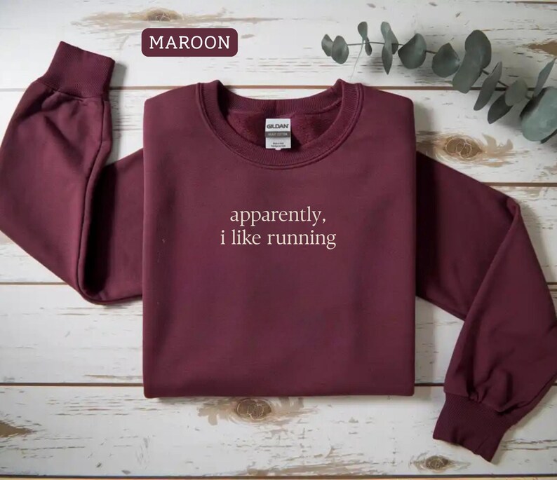 Może przedstawiać: Bordowa bluza z okrągłym dekoltem z białym napisem "apparently, i like running". Bluza ma długie rękawy ze ściągaczami i ściągaczem przy szyi. Słowo "MAROON" znajduje się w ramce w lewym g&oacute;rnym rogu.