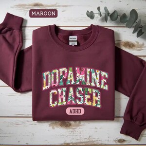 Pode incluir: Camisola bord&ocirc; com a frase "Dopamine Chaser" em um design floral. A palavra "ADHD" est&aacute; abaixo do texto. A camisola tem mangas compridas e gola redonda.