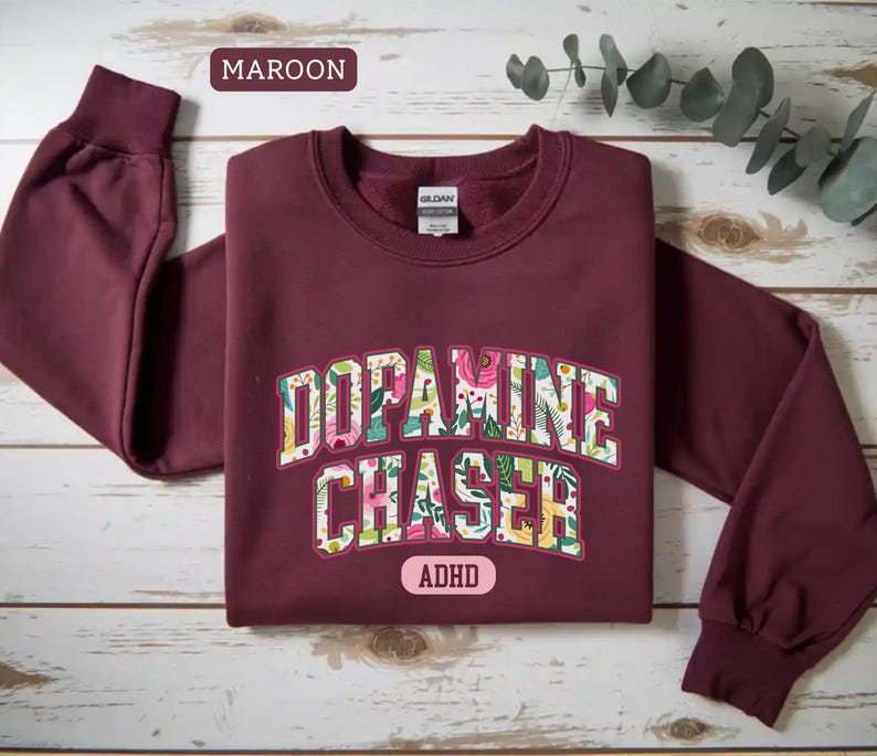 Pode incluir: Camisola bord&ocirc; com a frase "Dopamine Chaser" em um design floral. A palavra "ADHD" est&aacute; abaixo do texto. A camisola tem mangas compridas e gola redonda.