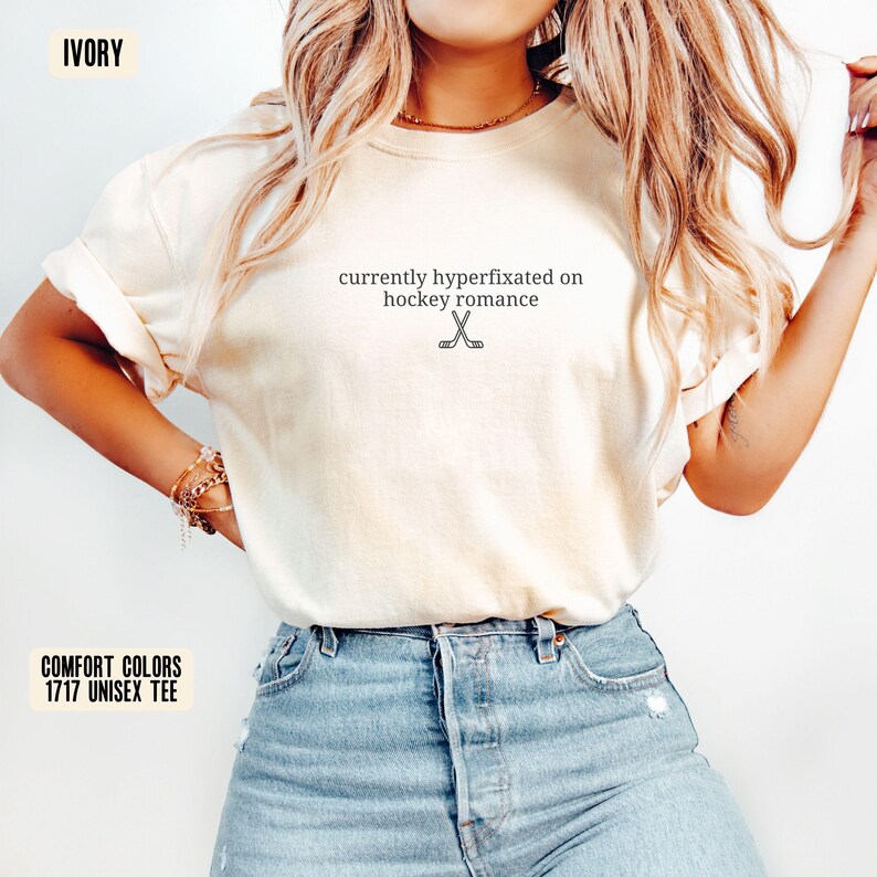 May include: An ivory short-sleeve t-shirt featuring the black text currently hyperfixated on hockey romance centered on the chest, accompanied by a small graphic of two crossed hockey sticks positioned directly below the printed text.